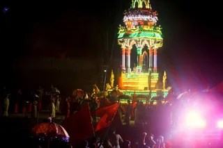 Night procession at a temple-style festival with a brightly illuminated ratha chariot in green, yellow and red lights, its tiered canopy topped with carved figures and sculpted horses at its base. Crowds of devotees in white and red-saffron clothing walk and dance around it, many holding red flags and carrying decorative umbrellas, with a strong pink-white stage light flaring from the lower right. The scene is a nighttime chariot procession in an open outdoor setting.