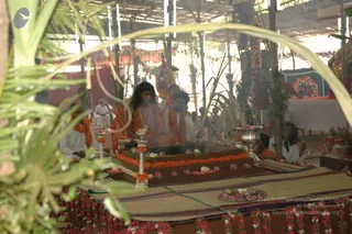 A photograph of a fire ritual (homa/yagna) in an open pavilion with a thatched roof and palm-frond decorations. In the centre, a long-haired man in orange robes with a flower garland sits behind a raised square fire pit, its edges lined with orange and white marigolds and pink blossoms. Smoke rises from the pit. Beside him, a man in white clothing sits at the fire, and further figures in white and saffron stand and sit behind. Brass vessels, a kalasha and fruit offerings are arranged on the platform, and a woven mat covers the floor in front. Swamiji (probable): the central seated figure in orange with long hair, garland and saffron robes, presiding at the fire.