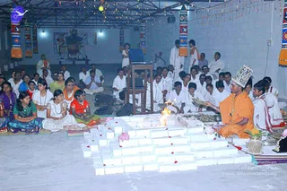 A homa is performed inside a hall with a white-tiled fire altar of stacked bricks at the centre, flames rising from it and rose petals scattered on the bricks. A man in saffron-orange robes and an ornate patterned crown sits at the right side of the altar, handling a plate of offerings and a ritual vessel. Around him, many people in white ritual clothes sit on the floor and stand, while women in saffron, red and green saris sit at the left. Behind the altar, a small deity murti stands on a table near strings of fairy lights and banners with blue and orange patterns hang on the pillars.