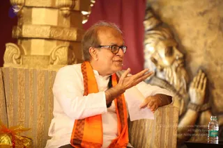 A photograph of a seated elderly man in a white kurta and dark-rimmed glasses, wearing an orange and brown checked shawl, gesturing with one raised hand while speaking on a striped upholstered sofa. Behind him stands a large gilded statue of a bearded sage with hands in anjali mudra, and a gold-and-cream carved pillar frames the left side. A water bottle sits on a dark table at right and a gold-wrapped gift is at lower left. A microphone clip is visible on his collar.