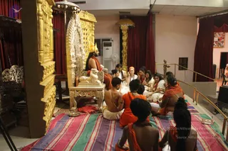 A photograph of a satsang in a hall with red drapes. A seated figure on an ornate golden throne with a lion-motif base wears a golden crown, long dark hair and saffron-orange cloth; he is the a person by appearance, presiding over the gathering. Around a striped multicoloured mat, devotees sit facing him: several women in white and cream clothes, a shirtless man in white dhoti, a woman in saffron robe, and two shaved-head sannyasis in orange in the foreground. A golden ornate palanquin-style canopy frames the throne, with a standing fan and air conditioner along the wall.