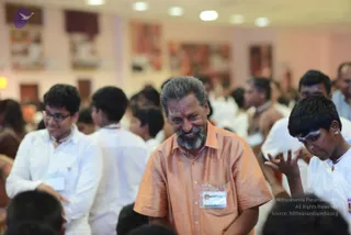 A photograph inside a bright hall with pale walls and framed artwork. In the centre, a smiling older man with grey hair and a grey beard wears a short-sleeved orange-peach shirt and a plastic name badge on a lanyard. To his right, a young boy with tilak markings on his forehead and a white shirt reaches out with his hand raised. Around them, many people in white shirts and white dhotis stand and sit, including a young man in glasses on the left, and others wearing lanyards.