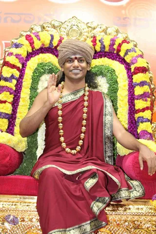A man seated on an ornate gold throne decorated with garlands of yellow marigolds, purple flowers and green leaves, raising one hand in greeting. He wears a brown turban, a red bindi on his forehead, a maroon robe with gold-patterned borders and a long gold bead necklace. Behind him is an orange backdrop with partial Telugu-script lettering. The setting suggests a formal satsang seat. Appearance-based candidate: Swamiji (probable), with long hair, a turban, saffron-toned ceremonial dress and gold beads seated on a throne.