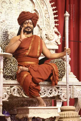 A man seated on an ornate silver throne with carved floral scrollwork and a leopard-print cushion, raising one hand in a blessing gesture and holding a tall silver staff in the other. He wears a rust-orange turban with a red-brown tilak on the forehead, an orange-saffron wrap with gold-bordered beaded trim, a gold waist belt, a gold chain, and a red thread on his wrist. Long dark hair falls behind his shoulders. Red draped curtains and a microphone stand appear behind him. a person: the saffron attire, long swept-back hair, ornate gold necklace and throne setting are consistent with the page's identification, but the appearance cues alone are not fully conclusive. The figure is seated on a throne in a stage setting.