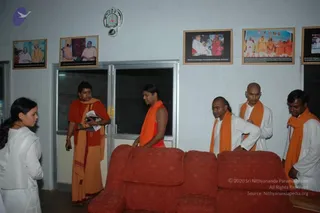 Indoor photo in a room with white walls and framed photographs, including a portrait of two saffron-robed figures and group photos with robed devotees. A woman in white stands at left, looking toward a man in a saffron-and-red robe holding papers and a folder. Beside him a man in saffron with an orange shawl turns toward the room, and two bald men in white garments with orange shawls stand near a red upholstered sofa. A man in white with an orange shawl stands at right. Glass doors are behind them.