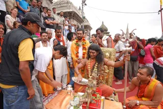 Outdoor temple scene at Pashupatinath with a crowd of onlookers behind a barrier. A figure with long dark swept-back hair, wearing orange-saffron cloth and a multi-strand gold-and-orange necklace, sits on a gilded golden throne-like seat and raises one hand over a small brass deity murti and offerings on a low table. A tall silver trident stands to the right. Nearby, a man in an orange robe and orange-and-yellow garland stands behind the seat, and a man in a white kurta leans forward to make offerings over a brass vessel. A shaved-headed man in orange robes and rudraksha beads kneels at the right edge beside the trident. Devotees in white, orange and pink clothing surround the scene.