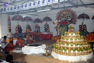 Photograph of a ritual hall with a white tiled wall and a row of Dhyanapeetam pennants overhead. Several deity murtis dressed in colourful garlands stand on draped platforms, with a large painted bull and a decorated arch behind them and red-and-orange parasols above. In the foreground, a priest in white dhoti and a man in orange robes with a rudraksha mala sit beside a square homa/yagna pit lined with white and red offerings. Mounds of brass-lidded pots decorated with mango leaves and white cloth sit on the right, and a brass oil lamp stands near the pit. Two men sit at the left with a drum and musicians behind them.