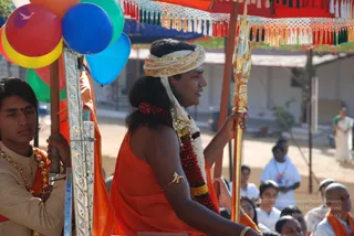 A procession scene outdoors under an orange and red fringed canopy. A central figure in saffron-orange robes, wearing a white and gold turban, a red and white flower garland, and a gold armlet, rides on a silver and gold palanquin-style ratha and holds a gold ceremonial staff. To the left, a young attendant in cream and gold ceremonial dress with a gold garland holds a bunch of colourful balloons tied to the ratha. Seated devotees in white and orange clothing watch from the background, and a figure in white stands at the right.