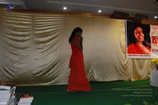 A person in a long orange-red garment with a shawl-like wrap stands barefoot on a green carpeted stage, palms pressed together in namaste. Behind them hangs a shimmering gold draped curtain. On the right, a large banner shows a smiling woman with long dark hair in orange clothing and a beaded necklace, labelled with Tamil text and "Paramahamsa Nithyananda". Stacks of orange-covered books sit at the left edge, and yellow flowers decorate a table at right.