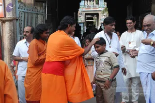A photograph of a temple entrance area with a gateway tower behind. A man in a saffron-orange dhoti and shawl, with long dark hair, places his hand on the head of a young boy in a striped t-shirt and khaki shorts, who wears a wristwatch. Around them stand men in white shirts and white dhotis; one wears a rudraksha-style mala, another has a sacred ash mark on his forehead, and a man in a light blue shirt holds up a small packet. A saffron-robed figure stands at the far left edge. The man in saffron has long, swept-back hair and a red cloth at his waist, and there is a Swamiji (probable) cue: long swept-back hair, saffron clothing and a shawl. Several other people, including a woman in a striped sari, are visible in the background crowd.