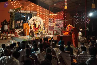 Night photograph of a temple-courtyard puja at the Sri Maheshwara Temple, lit by strings of warm lights. A large deity murti with a white halo-like backdrop and multiple arms stands behind a decorated platform heaped with fruit, brass vessels and lamps. A man in a saffron-orange dhoti and shawl stands at right, gesturing with a ritual object, while another figure in saffron sits on a throne-like seat near a shrine with a flower garland. Dozens of seated devotees in white dhotis and shirts fill the foreground, watching. A banner on the right shows photographs and text. The scene reads as a ritual worship gathering.
