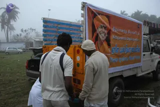 A photograph of a white relief vehicle with a large orange banner on its side showing a smiling man with an orange turban, a red tilak on his forehead and a long dark beard, beside the printed name Nithyananda Dhyanapeetam and the Tamil text Thane cyclone relief works. Three men stand at the rear of a yellow-and-blue striped truck, one in a white shirt with a dark bag strap, one in a cream sweater and white cap, and a third partly visible in white. Palm trees and a misty grey sky frame the scene on wet grass.