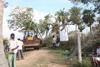 A photograph of a rural dirt road after storm damage, with fallen and broken trees and palms on both sides. A yellow JCB backhoe loader stands on the track, its operator visible in the cab. Several men in light shirts, lungis and sandals stand near the road; one in a white shirt and checked lungi holds papers beside a white cloth hung on a wooden pole. A wire mesh fence runs along the right, and a seated man in a white shirt writes on papers at the far right. Power lines cross the sky above.