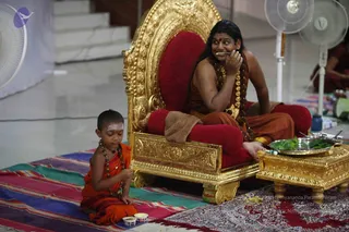 A photograph of a seated man on an ornate gold-and-red throne (peetha) in a bright hall, with a small boy in saffron-orange robes sitting on a striped rug beside him. The seated man has long dark hair, bare shoulders, a saffron-orange cloth, and a long rudraksha mala with a gold ornamental necklace; he rests his hand on his cheek and looks sideways. The boy wears an orange dhoti-style cloth, a beaded rudraksha-style mala, and a white tilak on his forehead, and is eating from a small cup. A silver tray of green leaves and food sits on a gold low table, and two pedestal fans stand nearby. Two people in red clothing are partly visible at the right edge.