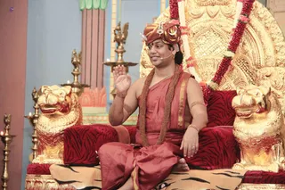 A photograph of a man seated cross-legged on an ornate golden throne with red tiger-striped cushions and golden lion-head armrests. He wears a rust-red turban with a jewelled ornament, a rust-red robe with gold border, a rudraksha mala and a long red-and-white flower garland draped over the throne. He raises one hand in greeting while speaking into a head microphone. Golden oil lamps stand beside him, and a golden lion sculpture sits at the left edge. Swamiji (probable) is seated in the centre, identified by the turban, rudraksha mala and saffron-red ceremonial robes, and presented on the throne as a teacher addressing a satsang.