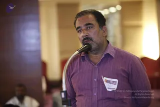 A man with short dark hair and a mustache stands at a microphone on a stand, speaking in a hotel-style hall with warm lights and red chairs. He wears a purple striped button-up shirt and has a white name badge pinned to his pocket. Blurred figures sit in the background at the lower left.