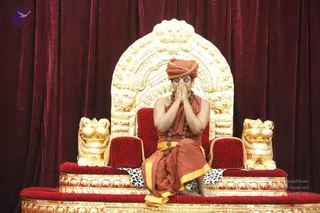 A seated man in an ornate golden throne with a carved arched backrest and lion-head armrests, set against deep red velvet drapery. He wears an orange-red turban with gold trim, an orange-red shawl over an orange-red dhoti with a gold border, and multiple rudraksha malas around his neck, with his bare shoulders and feet visible. He sits with palms pressed together in namaste and eyes closed, on a red-cushioned dais. No other people are visible. Swamiji (probable) is shown as the seated presenter, identified by the distinctive orange-red attire, rudraksha malas and turban on the throne.