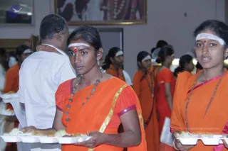 Indoor dining hall during a Ganesha festival meal. Two young women in the foreground wear orange saris with gold borders and white forehead bands with red bindis; one carries a white foam tray of food, the other holds one at the lower right. A man in a white shirt with a saffron-and-white shawl stands behind them, and more women in orange saris stand in the background. A framed portrait hangs on the wall above, showing a figure with garlands, but no one in the scene is identified as a person.