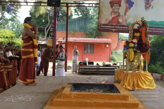 Outdoor ritual at a raised stone platform beside a square orange fire pit with a small flame and burning offerings. A man in a red-orange dhoti with a yellow border stands at left, hands folded in namaskar, wearing a long garland of red and yellow flowers. Behind him several priests in similar red and yellow dhotis hold silver trays. A woman in a white sari with yellow border stands at centre with hands folded, and another person in white stands nearby. To the right a golden elephant pedestal holds a seated figure draped in a red cloth and marigold garlands, beside a golden kalasha-like vessel and a red-and-gold canopy. Rose petals are scattered on the ground. A large banner behind shows a portrait labelled "Paramahamsa Nithyananda" wearing a red-orange turban, rudraksha mala and saffron clothing, with a world map beside it. Orange buildings and trees form the background.