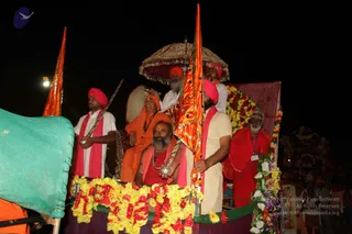 Night procession on an open decorated vehicle draped in yellow, orange and white marigold garlands, with a tall orange-saffron flag and a silver-trimmed brown umbrella overhead. A man in a white kurta and pink-red sash stands at the right holding the orange flag, and turbaned men in pink and orange turbans with white shirts stand around the platform. Seated at the front, a bearded man in a red robe with a rudraksha mala and a silver staff sits beside a woman in orange sadhvi clothing with a mala. A bearded elderly sadhu in red robes with an identity badge sits at the far right. Crowds and lit decorated floats are visible in the dark background, and a green netting covers the left edge.