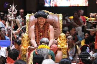 A seated figure in an ornate gold-and-brown throne flanked by golden lion heads sits at the centre of a crowded hall. He wears a dark swept-back hairstyle with a white flower, a long rudraksha mala and gold necklaces, and a red-orange wrap. A kneeling older woman in a green top with a yellow marigold garland bows at his feet, which he touches. Behind them, a large trident stands at left, a screen glows at top, and many seated attendees in orange and white clothing watch. A plate of offerings appears at right. a person is seated on the throne with long hair, rudraksha mala and gold necklaces, receiving devotion as a teacher.