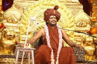 A seated man in a maroon-red turban with a small gold emblem and crescent ornament, wearing a maroon-red shawl over bare shoulders and long pink-and-white flower garlands. He sits on a leopard-print cushioned throne with gold lion-head armrests and an ornate gold backdrest, smiling and gesturing with his right hand toward a microphone on a stand. A white serpent-shaped staff rests at left, with a small framed photograph on a silver stand beside it. Swamiji (probable) appearance cues: maroon-red turban and shawl, ceremonial garlands and gold-throne setting, presented in a seated discourse role.