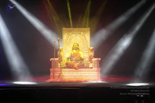 A golden statue of a seated figure sits on an ornate gold throne with carved lion-like armrests and a tall decorated backrest framed by a floral arch. The statue wears saffron-toned robes, a long rudraksha-style mala necklace and sits in meditation posture with hands joined. Two tall staffs or rods stand beside the throne on a stage lit by bright white and yellow spotlights beams in haze. The setting is a dark stage floor with a small purple bird logo at upper left. No living people are visible.