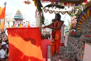 Outdoor photo of a festival procession on a decorated platform. A man with long swept-back dark hair wears an orange-saffron dhoti and a red-and-white flower garland, holding a large orange flag on a pole with both hands while smiling. Behind him a zebra-print sofa sits on a red-carpeted dais under a canopy of marigold and leaf garlands and coloured bunting. A crowd of onlookers, many in white, fills the street below, with orange flags, a multi-tiered coloured temple tower and a gopuram visible in the background. Probable Swamiji by appearance cues: long hair, saffron clothing and garlands on a ceremonial platform.