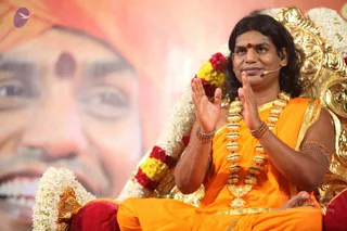 A seated man in saffron-orange robes sits on an ornate gold throne decorated with white jasmine garlands and red, yellow and white flower strands. He wears long swept-back dark hair, a red bindi on his forehead, a head-mounted microphone, a heavy gold-toned necklace and rudraksha-style beaded bracelets, and he presses his palms together in a gesture of greeting. Behind him a large blurred portrait shows a smiling face with a red tilak, and a saffron backdrop fills the background. a person seated in satsang.