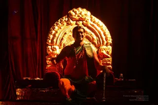 A man seated on an ornate golden throne with a large arched gilded backrest against deep red velvet drapery. He wears an orange-red saffron wrap over his torso, a green and gold bordered cloth at the waist, and a long rudraksha mala with ceremonial gold details. His right hand is extended forward and his left rests on a stick or staff. A small bottle and draped cloth sit on the stage ledge. The lighting is warm and dramatic.