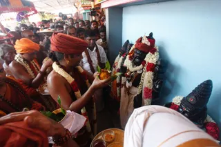 A crowded temple-style ritual scene. A bare-chested priest in a rust-red turban and dark red garland holds a coconut with green leaves, offering it toward black stone deity murtis adorned with white jasmine and red flower garlands, one with a silver crown. Nearby, a man in an orange turban and white-beaded garland stands beside a shaved-headed devotee in saffron. A man in a white shirt with a saffron shawl watches, a brass plate with kumkum and offerings rests in the foreground, and a large crowd presses behind, with a camera operator visible at upper right. Blue wall and tent canopy frame the scene.