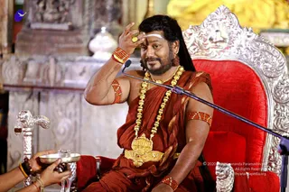 A bearded man in rust-red saffron-toned robes sits on an ornate silver-and-red velvet throne, touching his forehead with his right hand, which wears a gold ring and red beaded bracelets. He has white vertical tilak and a red bindi, a heavy gold necklace with a large ornate pendant, and a gold-and-red waist belt. A microphone on a stand stands before him. At lower left, a hand holds out a silver bowl near a silver-handled object. Behind him are carved stone pillars and gold-and-yellow temple decoration.