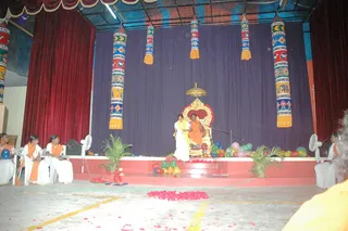 A stage event in a hall with a dark blue backdrop and red side curtains, decorated with tall painted cylindrical banners in red, blue, yellow and white patterns. A figure in orange robes sits on an ornate gold throne with a small gold canopy, a red cushion behind it. Beside the throne a woman in a white sari with a yellow shawl stands near the stage edge. Multicoloured balloons, potted palms and scattered red petals decorate the platform, and a few seated women in white with orange shawls sit at the side of the stage.