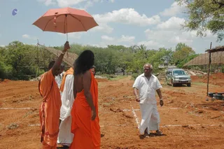 Outdoor photograph of a construction site at a red-earth plot with bamboo-framed structures and trees. A long-haired figure in an orange saffron garment stands centre-left, bare-shouldered with the orange cloth draped over the body, while another orange-robed person on the left holds a pink umbrella overhead. A man in white shirt and white dhoti walks on the right toward a silver SUV parked behind. Survey lines marked in white on the ground and stacked tyres lie to the right. Appearance cues for the long-haired figure are partial, so identity is uncertain.
