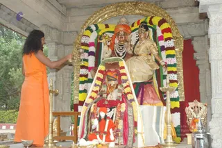 A figure in a long saffron-orange robe stands at left, back partly turned, reaching toward a brass oil lamp held before a shrine. Behind them a large deity murti pair sits under a gilded arch wrapped in garlands of yellow, white and multicoloured flowers: a male figure with an orange turban and layered white and black beaded necklaces, and a female figure in a cream and gold sari with a crown. A small orange-clad figure sits at the front of the altar, garlanded. Brass oil lamps stand on both sides and a silver vessel sits at right. The setting is a stone temple-style pavilion with carved pillars and greenery behind. No physical people beyond the robed figure are visible.