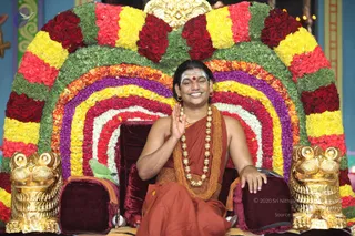 A seated man in rust-orange sannyasi robes waves his right hand in greeting while smiling from a velvet throne. He wears a long ornate golden necklace with beaded strands, sandalwood and white vibhuti marks on his forehead and a red kumkum bindu. The throne is backed by a large semicircular arch of fresh flowers in red, yellow, orange, purple and white, with golden lion-shaped armrest finials on each side. A small green parrot perches on the left armrest. Swamiji (probable), a candidate with long swept-back hair, saffron ceremonial clothing and gold necklaces, is seated on the throne as the central figure.