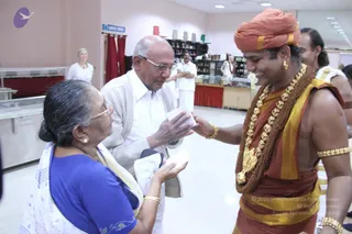 Indoor photo in a bright hall with pale pink walls, a white tiled floor, and a glass display case on the left. A standing man in a white shirt and cream cardigan with glasses hands a small white packet to a woman in a blue blouse and white saree with gold-embroidered border, who holds out her hands to receive it. Beside them, a smiling man wears an orange-red turban, ornate gold necklaces with a large brown rudraksha-style mala, gold armbands and earrings, and a red-gold dhoti; he is bare-chested with a gold-bordered cloth draped over one shoulder. Behind them, people in white clothes stand near a counter with shelves. Other people are partly visible at the edges and in the background, and a blue sign reads LADIES TOILET.