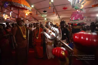 A photograph inside a festive tent decorated with hanging colourful tassels and lamps. In the foreground a large silver trident with white markings is carried, and a decorated kalasha topped with pink flowers and red garlands rises at right. A man on the left wears a rust-orange robe, a matching orange turban, a gold-coloured garland and a gold pendant; he smiles while standing among the crowd. Beside him a younger man in an orange robe wears a rudraksha mala and gold pendant. A man in white dhoti and gold-trimmed cloth stands near the trident, and a man in a dark shirt with a white dhoti walks at centre. Several devotees and onlookers fill the background, some in red and orange saris.