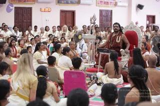 A large indoor hall with pale pink walls and wooden doors, where a crowd of many seated participants fills the floor. A man with long swept-back dark hair, wearing a rudraksha mala and a large ceremonial white and gold necklace over rust-red and saffron clothing, sits on a silver-framed red throne at the front right. Beside him stands a silver trident and a small silver fan stand, and a digital clock on the wall reads 06:05. Many participants wear white and cream traditional dress, some with rudraksha malas, and several sit on pink mats. Swamiji (probable) cues: long swept-back hair, rudraksha mala, ceremonial necklace and saffron clothing, seated on the throne as the teacher. Some people in the foreground hold pink name placards.