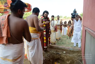 Outdoor bhumi puja at a construction site beside a red-washed wall. A bare-chested priest in an orange-saffron cloth and white dhoti stands at left with a yellow-bordered sash. Centre, a bearded man in a rust-brown robe and rudraksha mala wears a multicolour flower garland and holds a small object while standing at the edge of a freshly dug pit. Two men in white dhotis with yellow borders stand behind him, and a cameraman in white shirt and trousers films from right. Other figures in white and saffron wait in the grass, and an ornate fringed canopy appears at upper left. Petals lie in the dirt of the pit.