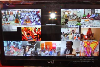 A photograph of a flat-screen TV displaying a video-conference grid of satsang participants from multiple locations, labelled Hyderabad Aadheenam, Selayang Malaysia, Karlsruhe Germany, Seremban Sivakasi, Dubai Adheenam, Philadelphia, Playa del Carmen Mexico, Melbourne Australia, Bangalore North, and others. Several panels show groups seated in white and saffron clothing around decorated altars with kalasha, flowers, oil lamps and fruit offerings, while other panels show a woman in red and gold, a man in a white shirt smiling, and a figure in a red and gold ornate seat.