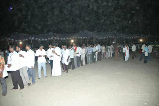 A night-time photograph of a long, orderly queue of men standing in a sandy open ground beneath a dark tent roof strung with small pennant flags and hung with lamps. Most men wear white shirts and white dhotis or trousers, while others wear checked or striped shirts. A few men at the far right wear saffron-orange cloth and move among the crowd near a parked green vehicle. The line stretches from left to right across the frame, with people standing close together facing forward.