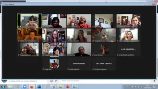 A screenshot of a Zoom Meeting gallery view showing sixteen video tiles of participants joining an online daily update session. Most tiles show women in home settings with framed pictures, shelves and deity images behind them. One tile shows a man in a dark shirt with a plain background. One tile shows a woman in a red and gold garment beside a figure in ornate red dress, and another shows a woman seated before a colourful deity image with a garland. Several tiles are text-only name labels with no video. The Windows taskbar and a chat bar appear at the bottom of the screen.