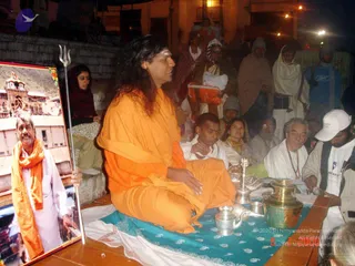 Evening outdoor ritual on a stone platform at night. A long-haired figure in saffron-orange robes and an orange shawl sits cross-legged on a teal and white cloth, facing left, with a trident (trishul) standing beside them. Brass and silver ritual vessels, a lamp and a small bowl sit on the cloth. A framed photograph on the left shows a white-clad elderly man with an orange shawl in front of a temple. Behind, a crowd of devotees in shawls, woollen caps and white clothing watches, including a man in a white cap and a bearded man in glasses. Illuminated buildings line the background. Swamiji candidate by appearance: Swamiji (probable), with long hair and saffron garments, seated in a ritual setting.