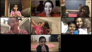 A screenshot of an online video call grid showing eight tiles of children and adults, most with palms pressed together in namaste. Top-center tile shows a smiling woman with a white forehead mark and orange top, labelled Nithyananda Vancouver, in front of a framed picture of a sannyasi in saffron robes with garland, flowers and a small altar. Tiles labelled Mythreyi, Janarajh K, Sherry B, Maya & Varun / Neha and Sarvesh show children indoors; one girl in pink hugs a woman with long dreadlocked hair in a pink shirt, and a boy in pink yoga mat setting stands with hands joined. Tile labels and call interface are visible.