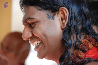 Close side profile photograph of a smiling man with long, wet, wavy dark hair, water droplets on his face, and a broad open-mouthed laugh. He wears a red garment and a large necklace of brown rudraksha beads. A blurred older person with a bald head sits in the background at left. The corner has a small purple bird logo and a , , Source