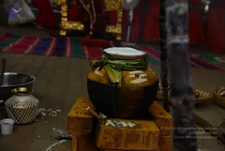 A black clay pot called a pongal pot stands on a stack of orange-brown bricks on a floor, decorated with white horizontal lines and a red dot, with green leaves tied around its neck and a white cloth or lid on top. Brass vessels and a woven basket sit nearby, with scattered white rice grains on the ground and a kolam line drawn on the floor. Behind, red-clad figures and a colourful striped mat appear softly out of focus, with gold-covered seats visible. No people are clearly in the foreground.
