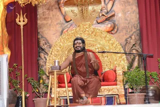 A man seated cross-legged on an ornate gold throne with a carved gold cobra hood canopy, speaking into a stand microphone during a discourse. He wears a rust-orange robe, has long swept-back dark hair, a red bindi, and long rudraksha mala necklaces hanging to his lap. His right hand is raised in an open gesture while his left holds a small metal cup on a wooden stool. A gold trident stands at left, potted plants flank the stage, and a large carved relief panel with bearded sage figures forms the backdrop beside red drapes. a person: the long swept-back hair, rudraksha malas, orange robe and ornate throne are the identifying cues.