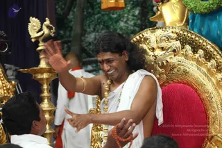 A smiling man with long dark hair and a red bindi sits on an ornate gold throne with a red cushion, waving his right hand while his left hand is extended downward. He wears a white dhoti-style shawl over one shoulder, a long gold-beaded necklace of large gold bead strands, and a gold bangle. In the foreground a seated man in white leans in at the left, and a man in white with an orange-red cloth stands behind. A tall gold oil lamp with a peacock-shaped top stands at left, and a gold and green garlanded deity murti appears at upper right. a person cues: long swept hair, ceremonial gold necklace and white shawl, seated on a throne; the appearance match is strong but the face is not used for identification.