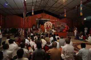 A photograph of an evening ritual in an open-sided shed with corrugated roof, strung red fairy lights and tall painted cloth banners hanging from the rafters. A large deity murti with a white halo-like arch stands on an altar decorated with flowers and oil lamps, with sign reading SRI KAILASESHWARA TEMPLE above. Several rows of seated devotees, many in white dhotis and shirts, sit on the floor facing the altar; a man in white stands at the altar tending the rites. On the right, a figure in orange robes sits on a raised red seat beside other saffron-clad attendants, and a standing man in white watches. A cameraman stands at the left edge. A sand-coloured circular mandala is visible at the lower right.